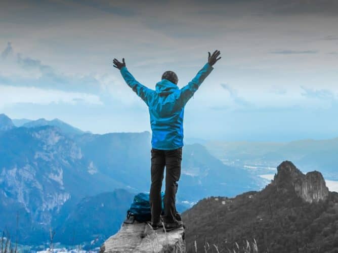 leveraging-vs-delegation-blog-man-arms-raised-open-standing-on-top-of-mountain-overlooking-view-thinking-without-restrictions-2023-1920×549 Man with both arms raised standing on top of a mountain